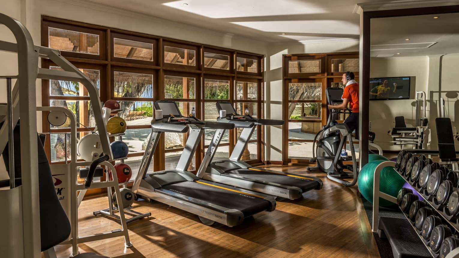 Gym with wooden floors, two treadmills, person on elliptical, free wights, windows looking out to beach