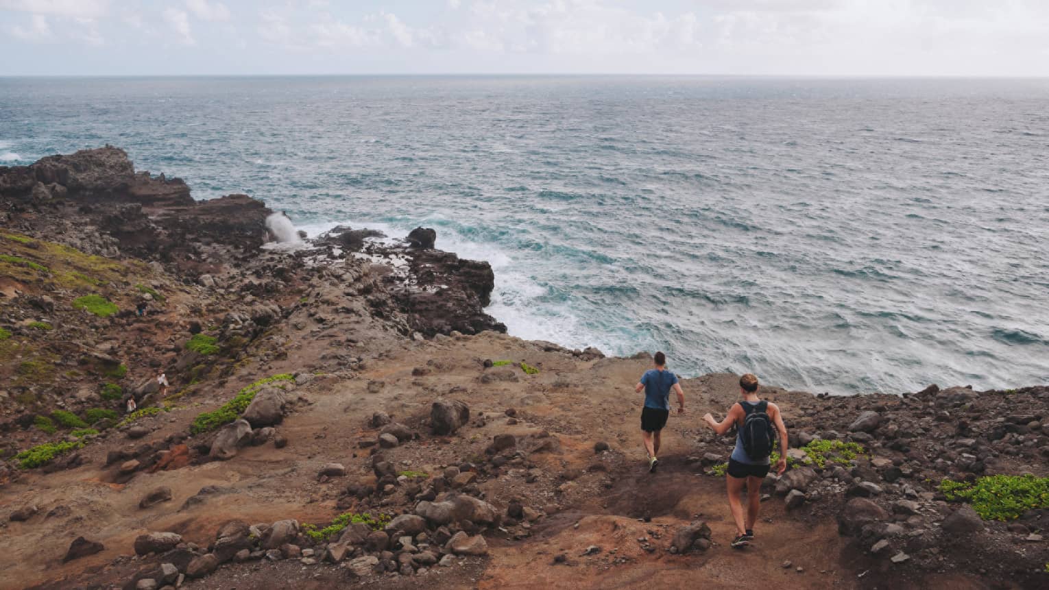 Two people hike down dirt-covered path, rocks towards ocean