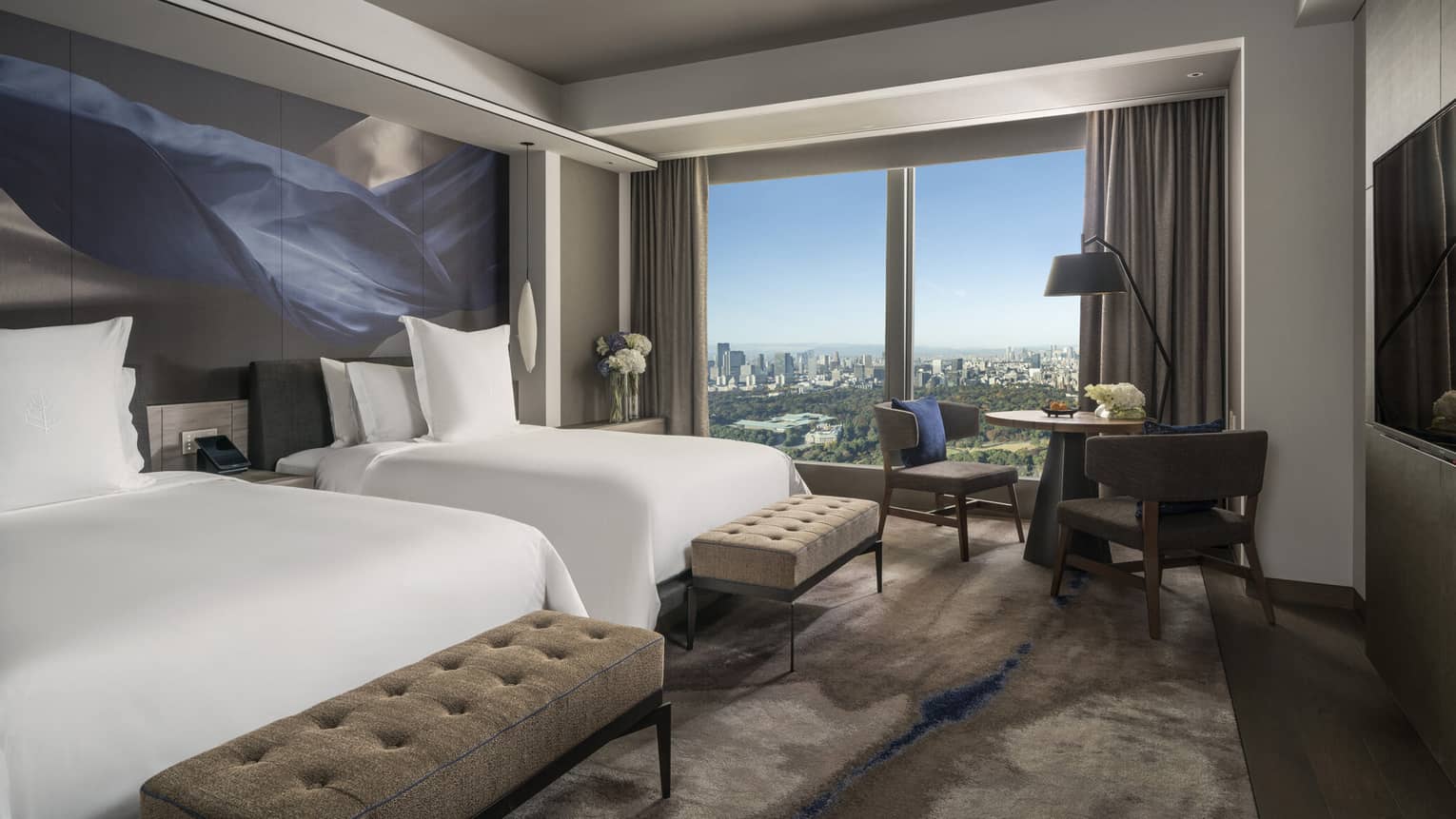 Guest room with two beds, tufted benches, seating area and floor-to-ceiling windows offering city views