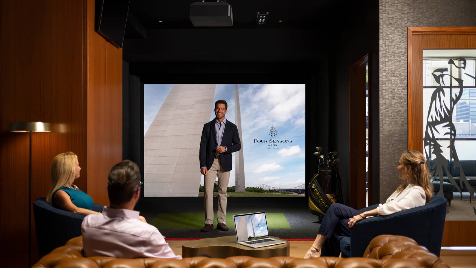 A man in a suit standing in a virtual golf room talking to three people sitting in chairs.