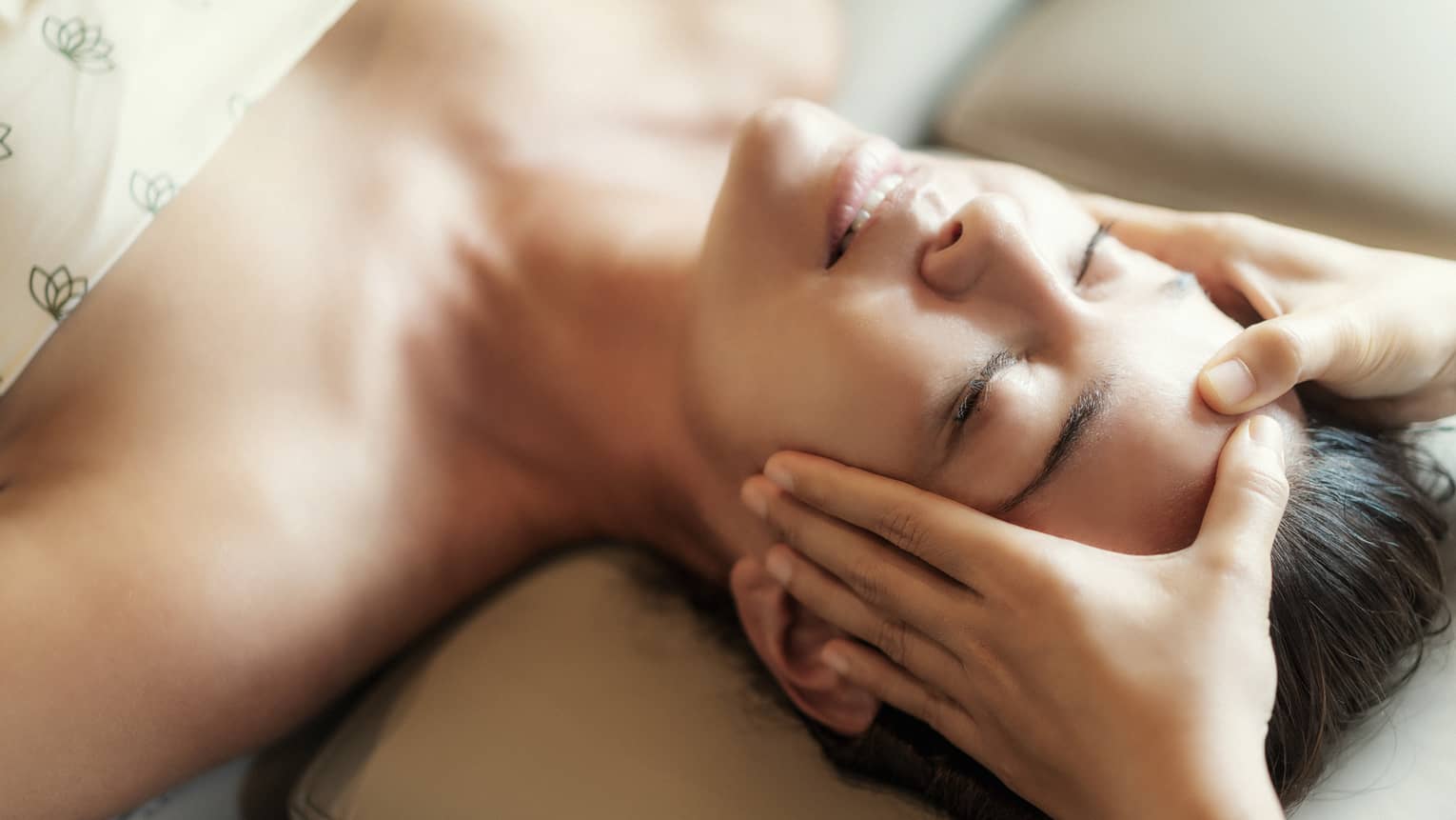 Spa facial, two hands rest on woman's forehead as she closes her eyes, lays under sheet on treatment table