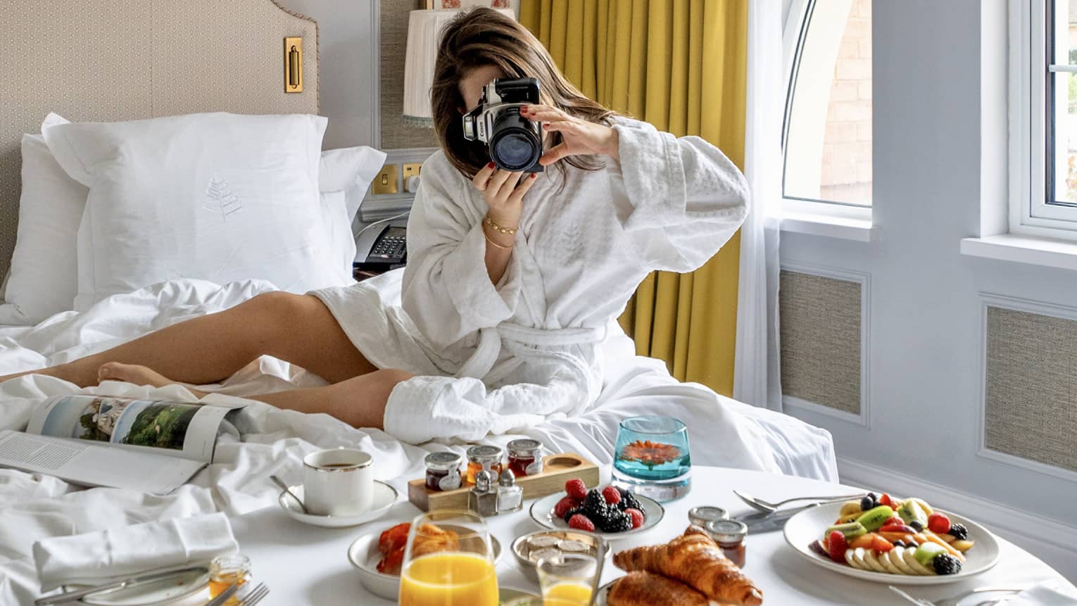 Woman in white robe sitting on hotel bed taking picture of In-Room dining breakfast spread before her