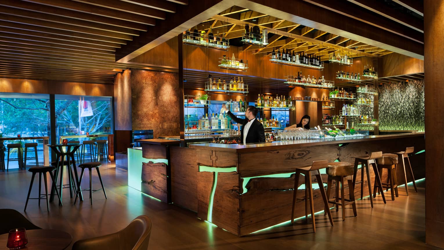 Grain bartenders reach for bottles, prepare drinks at rustic wood bar with lights