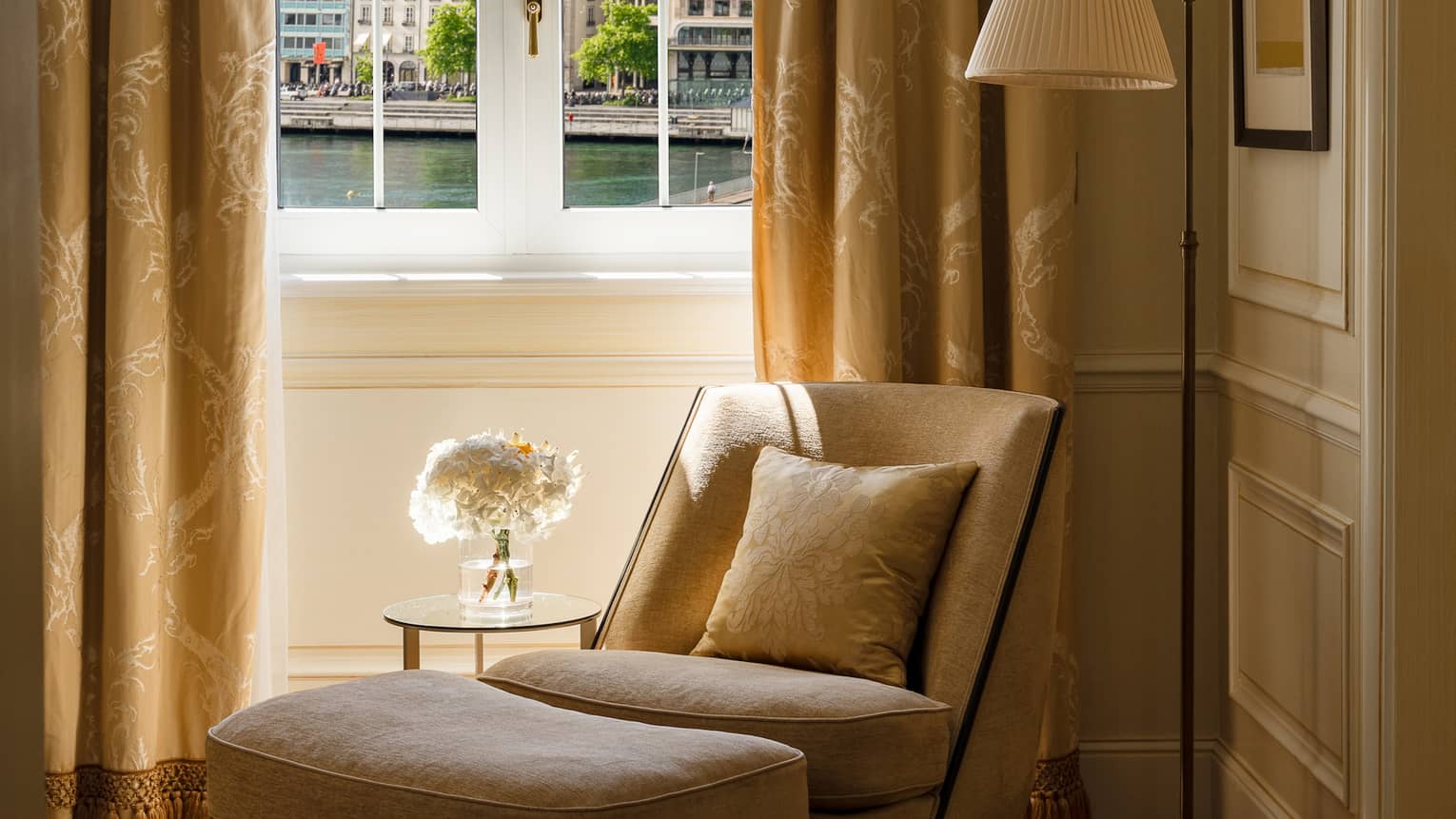 Luxury suite corner with a beige armchair with a matching ottoman next to a side table holding a floral arrangement, positioned by a window overlooking water