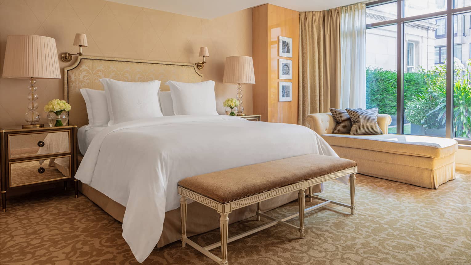 White bed with tan bench at foot against tan wall and chaise longue by large window
