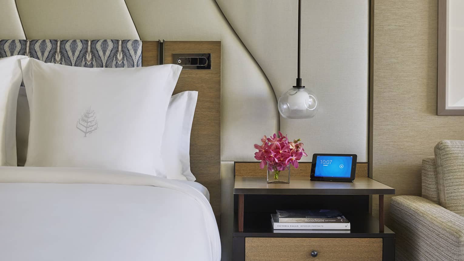 Hotel room with white bed, Four Seasons logo on pillow, large white padded headboard, flowers and ipad on nightstand