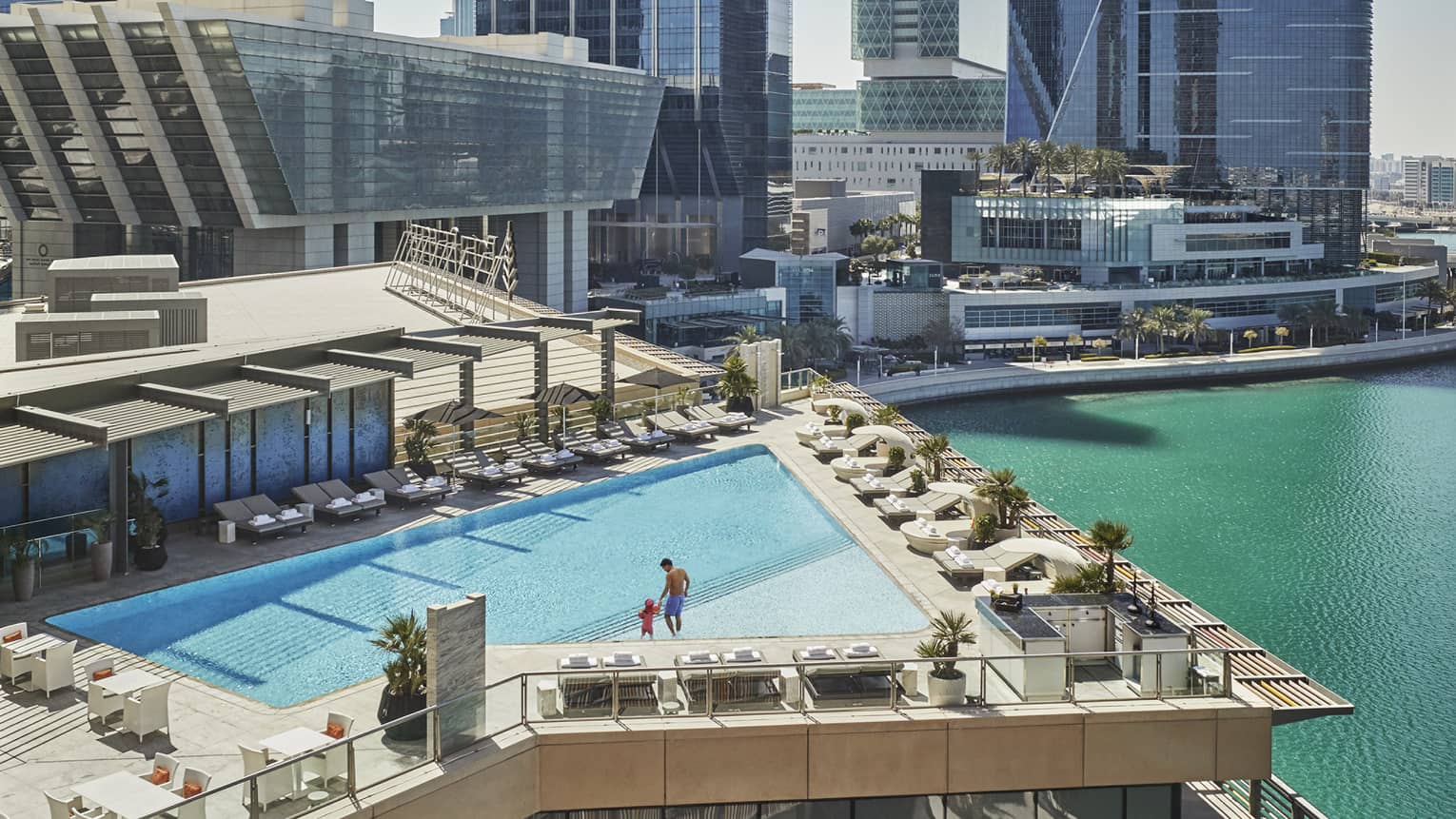 A wide shot of a man and child walking around a triangular pool overlooking the water and Abu Dhabi skyscrapers in the background. 