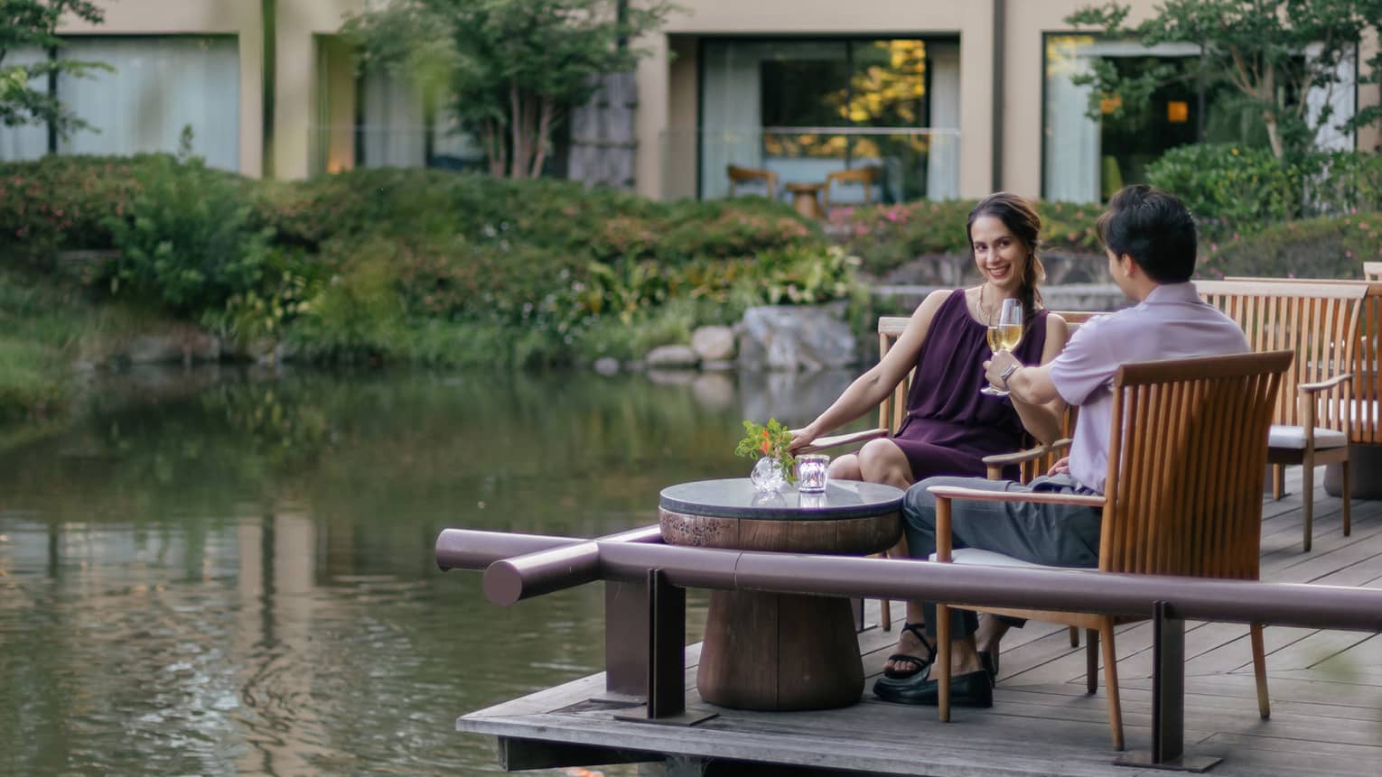 Smiling couple clink champagne flutes at a café table on a wood deck overlooking a rippling pond bordered by lush foliage.