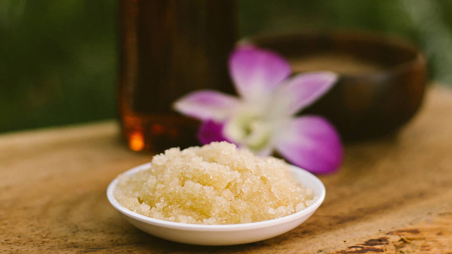 Bowl with salt scrub next to purple tropical flower and glass oil bottle on rustic table