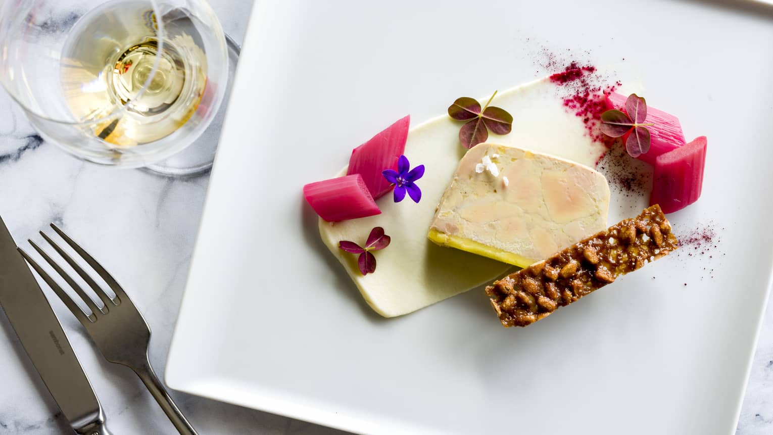 Foie Gras Gerbeaud with flower garnish on white square plate, knif & fork, white wine