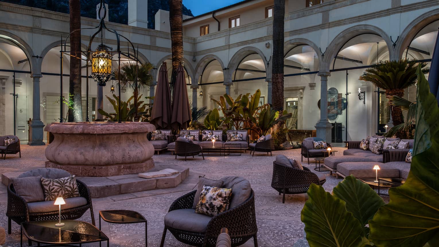 Outdoor hotel courtyard at dusk featuring chairs, tables and a central fountain, creating a serene lounge atmosphere