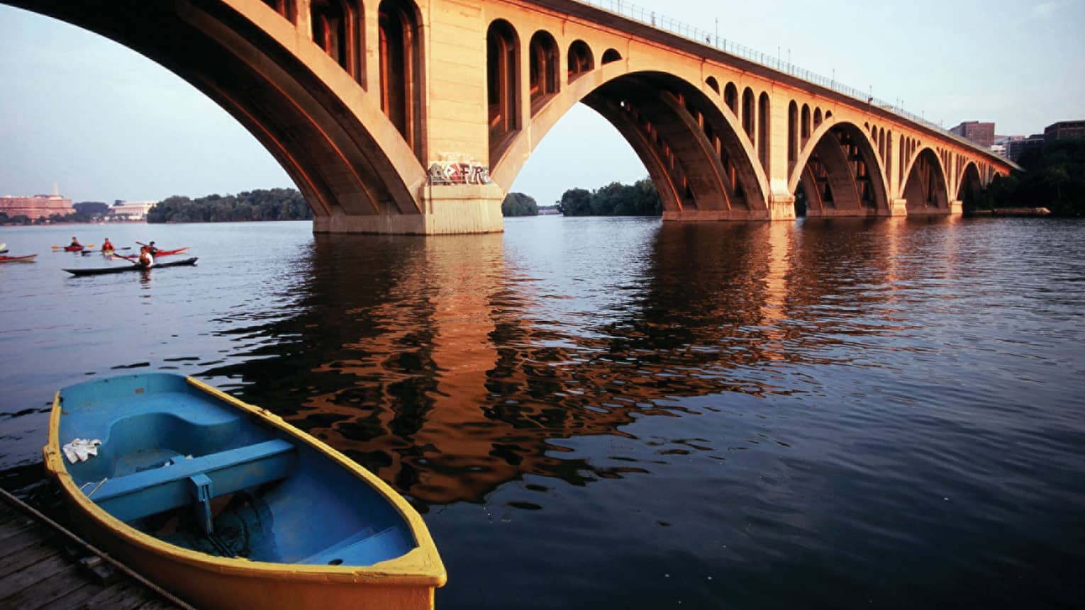 A wide river with a large bridge crossing it, there are people rowing kayaks under the bridge.