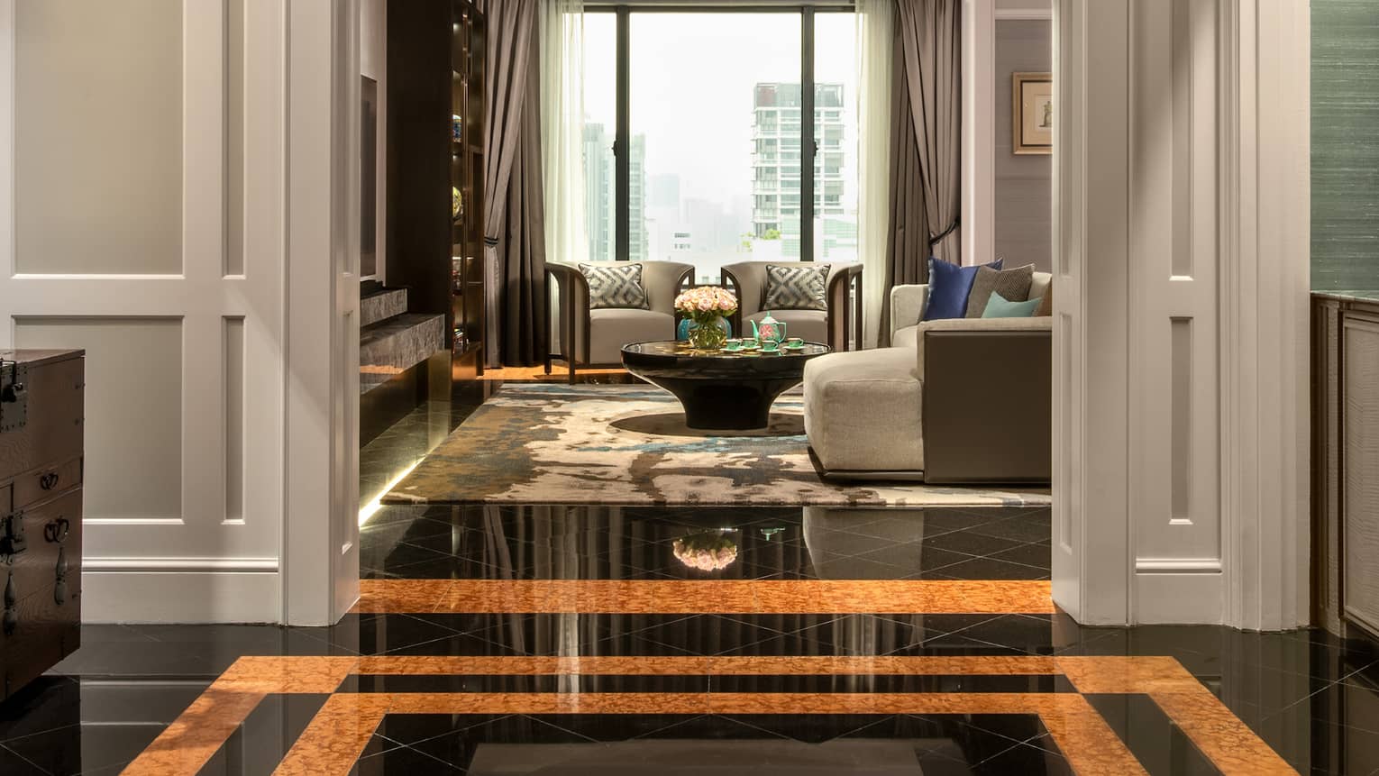 A hallway with glossy black and gold marble floors leading to a well-lit living area with modern furniture, large windows and a cosy seating arrangement.