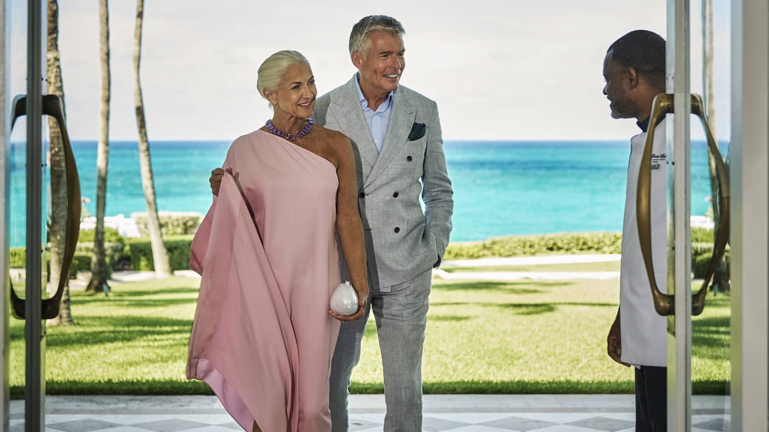 Woman in pink dress and man in gray suit walking through the doors with the ocean in the background.