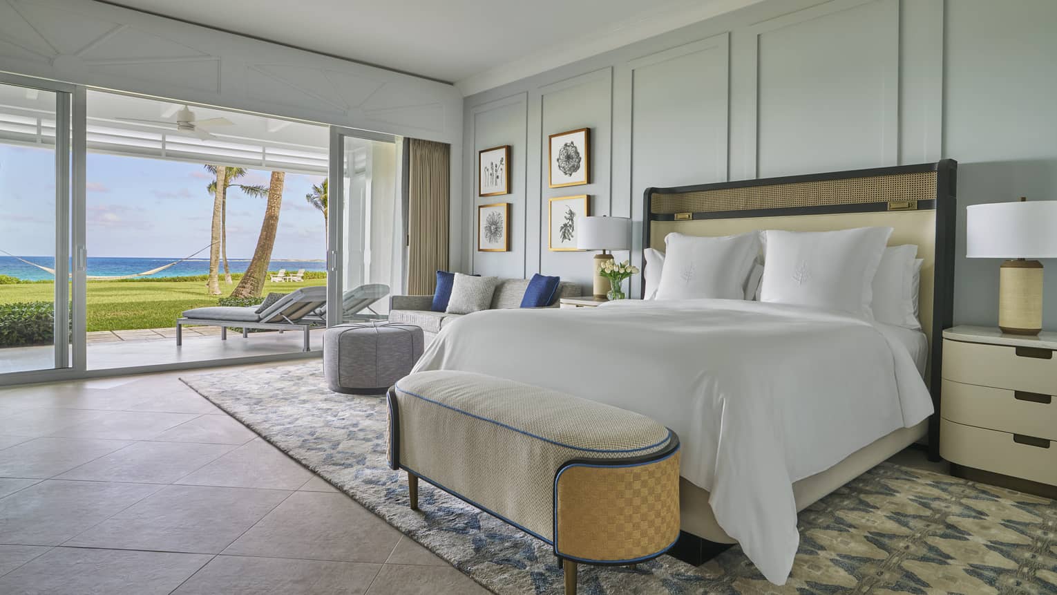 A guestroom with large windows looking out towards a beach, a bed with white sheets, pastel green walls and two nightstands with lamps.