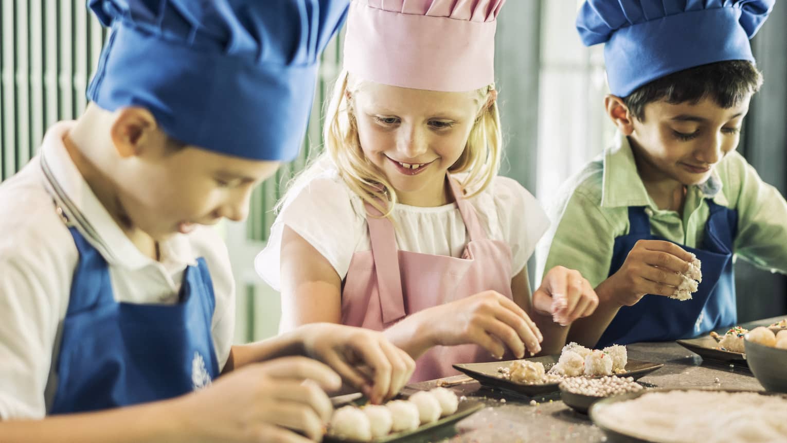 Three children in chef hats, aprons roll pastries at counter
