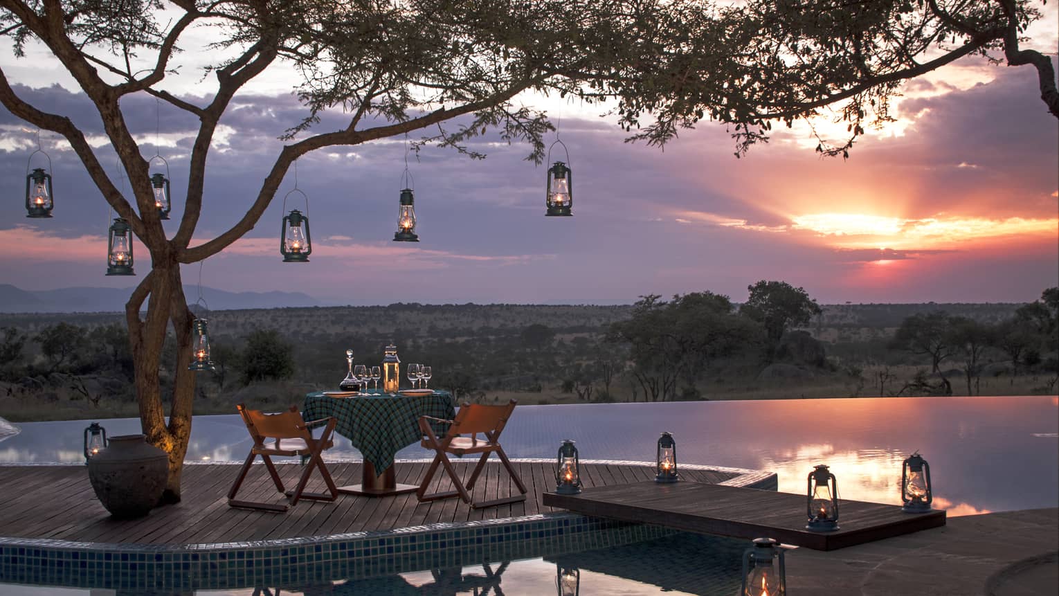 Private dining table on wood dock lined with lanterns, hanging from tree, sunset