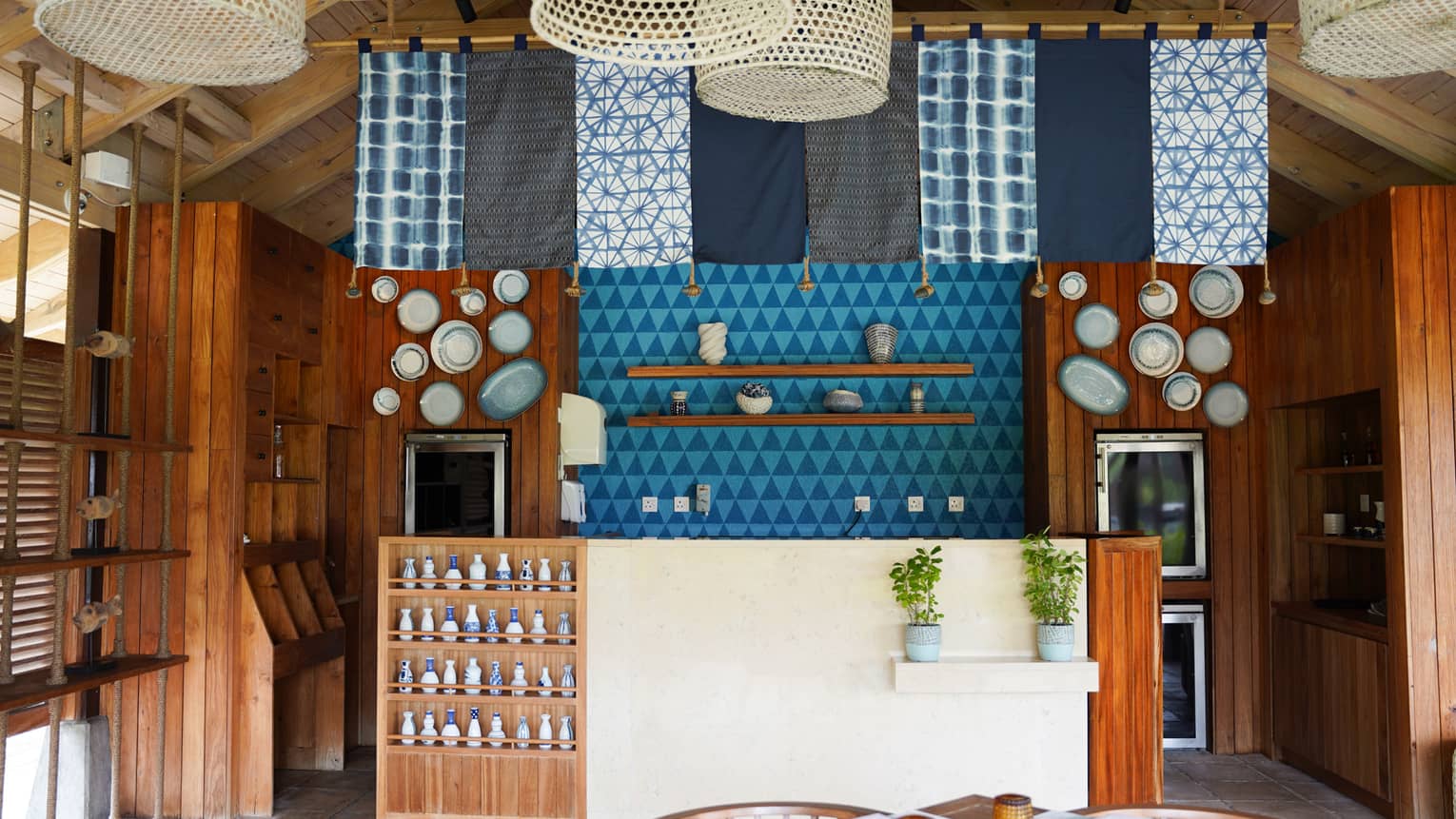 Casual restaurant with an island vibe, with blue and white fabric panels and rattan lights hanging from ceiling over teak and stucco bar