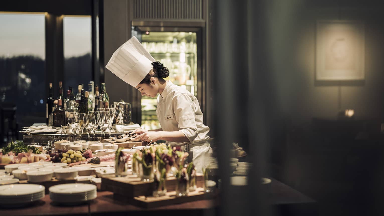Chef wearing white uniform, hat stands garnishes appetizers on counter covered with dishes, meals