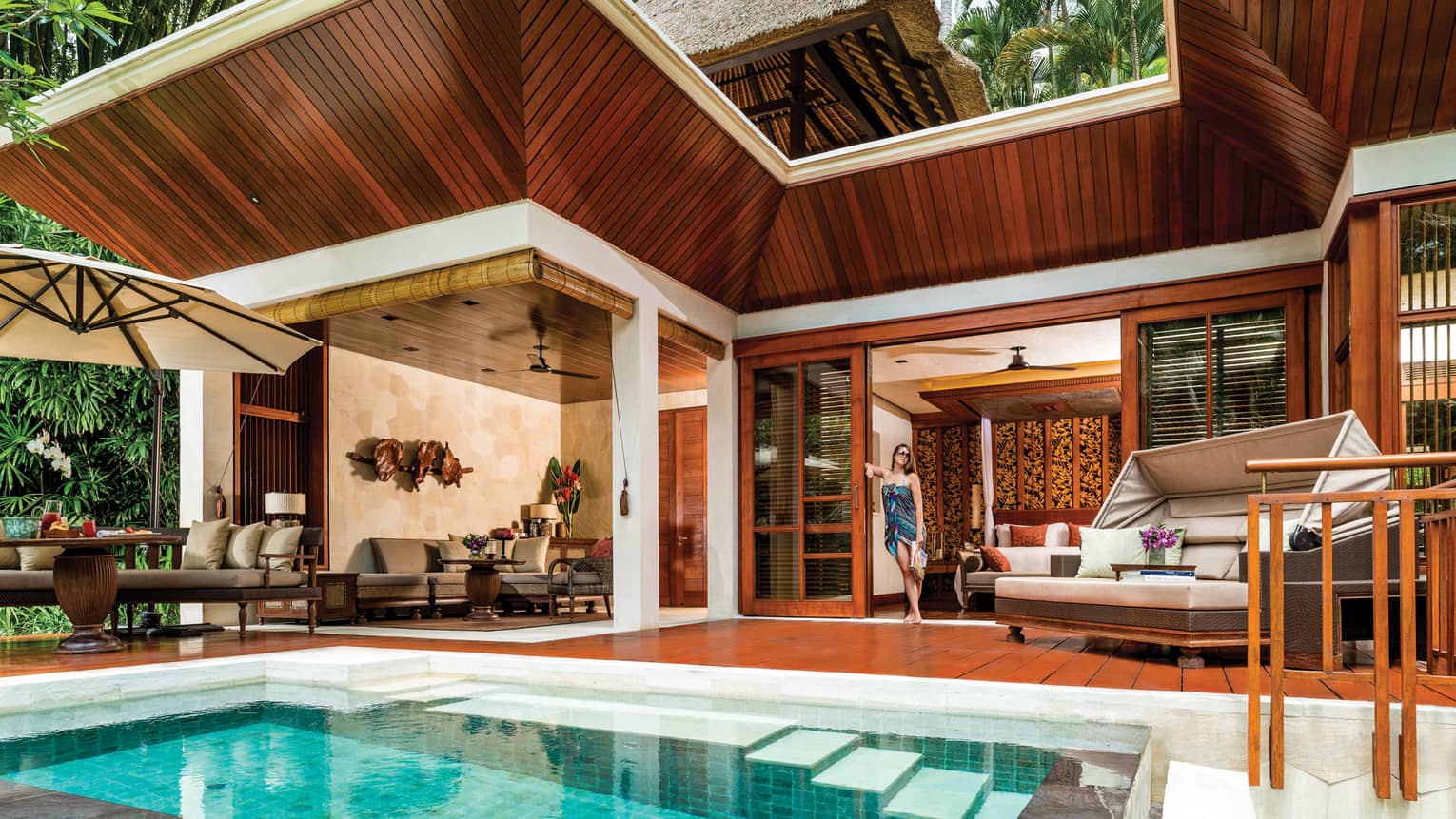 Woman in colourful swim wrap and sunglasses leans against wall of modern One-Bedroom Villa wood deck, by pool