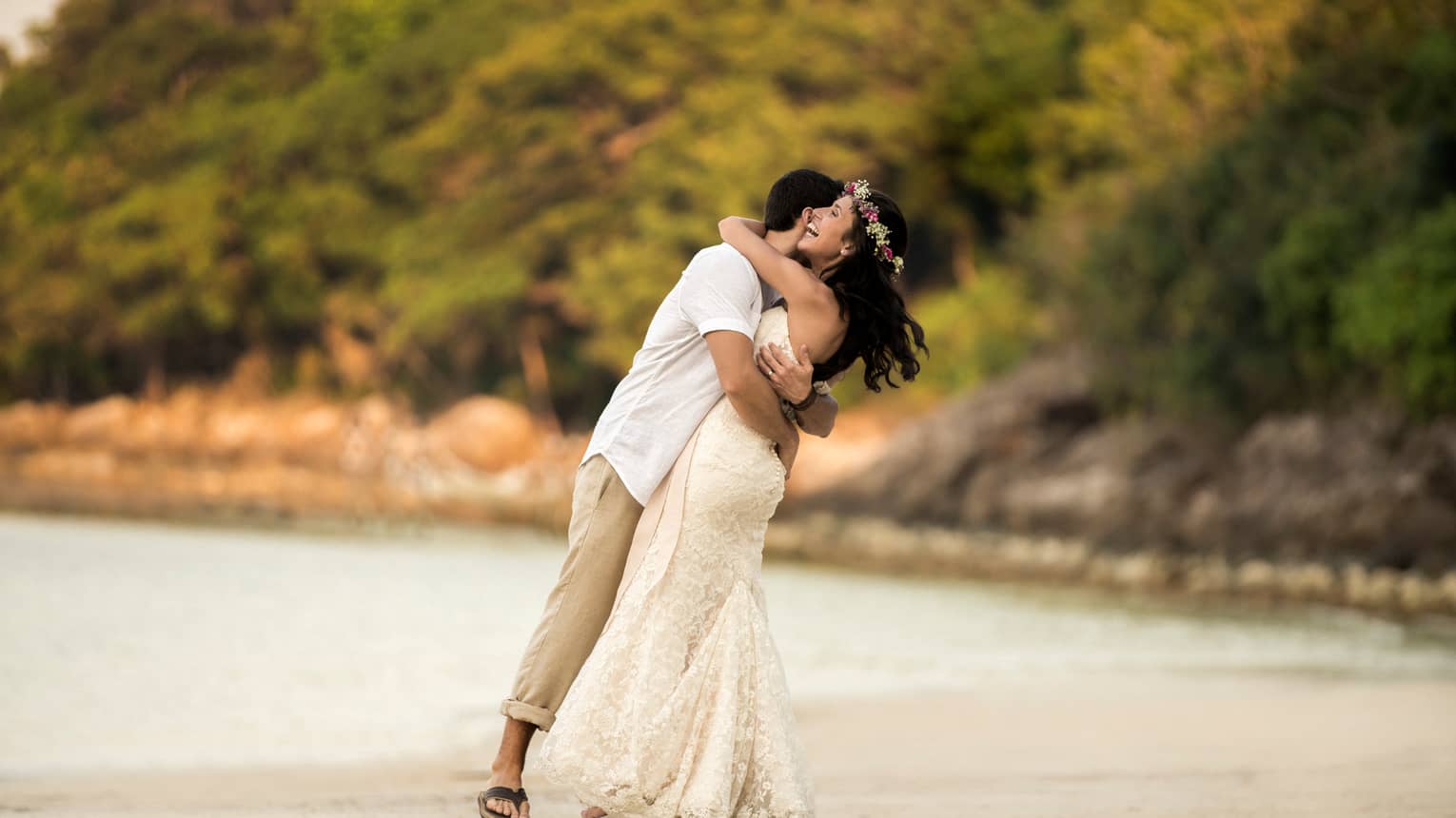 Man in white shirt and khaki pants, woman in long white wedding gown and flower crown embrace on the beach