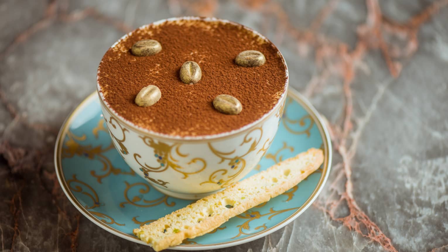 A cup of tiramisu topped with biscotti, served in a blue and gold teacup.