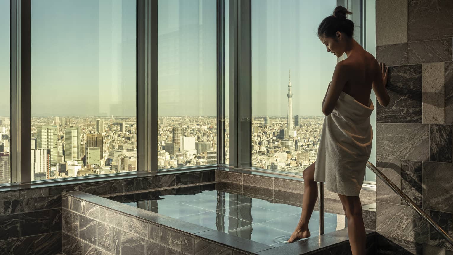 Woman wrapped in white towel dips her toe into the ofuro bath