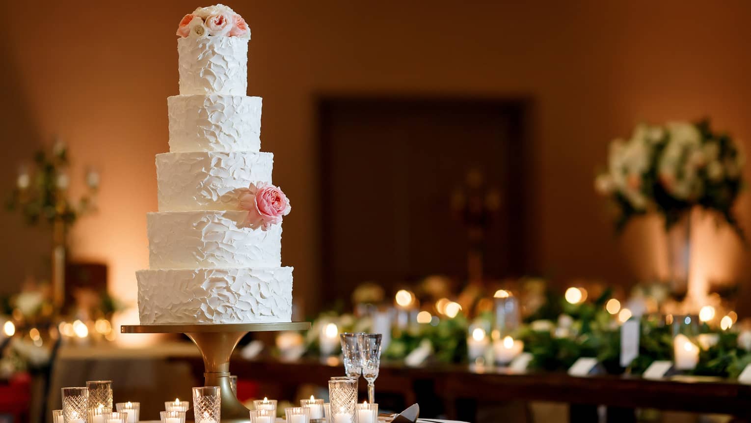 A five-tiered white wedding cake garnished with a white rose and some small white and pink flowers on the top tier , sits on a white round table surrounded by white candles