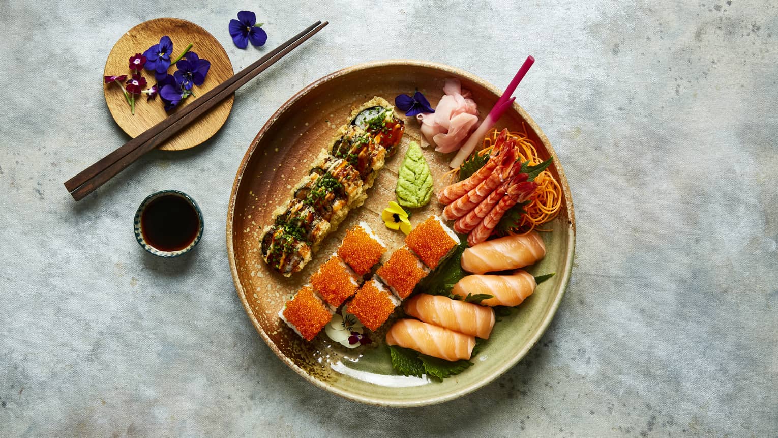 A ceramic bowl holding sushi and sashimi rolls, a small container of soy sauce, a small ceramic plate holding chopsticks and small purple flowers