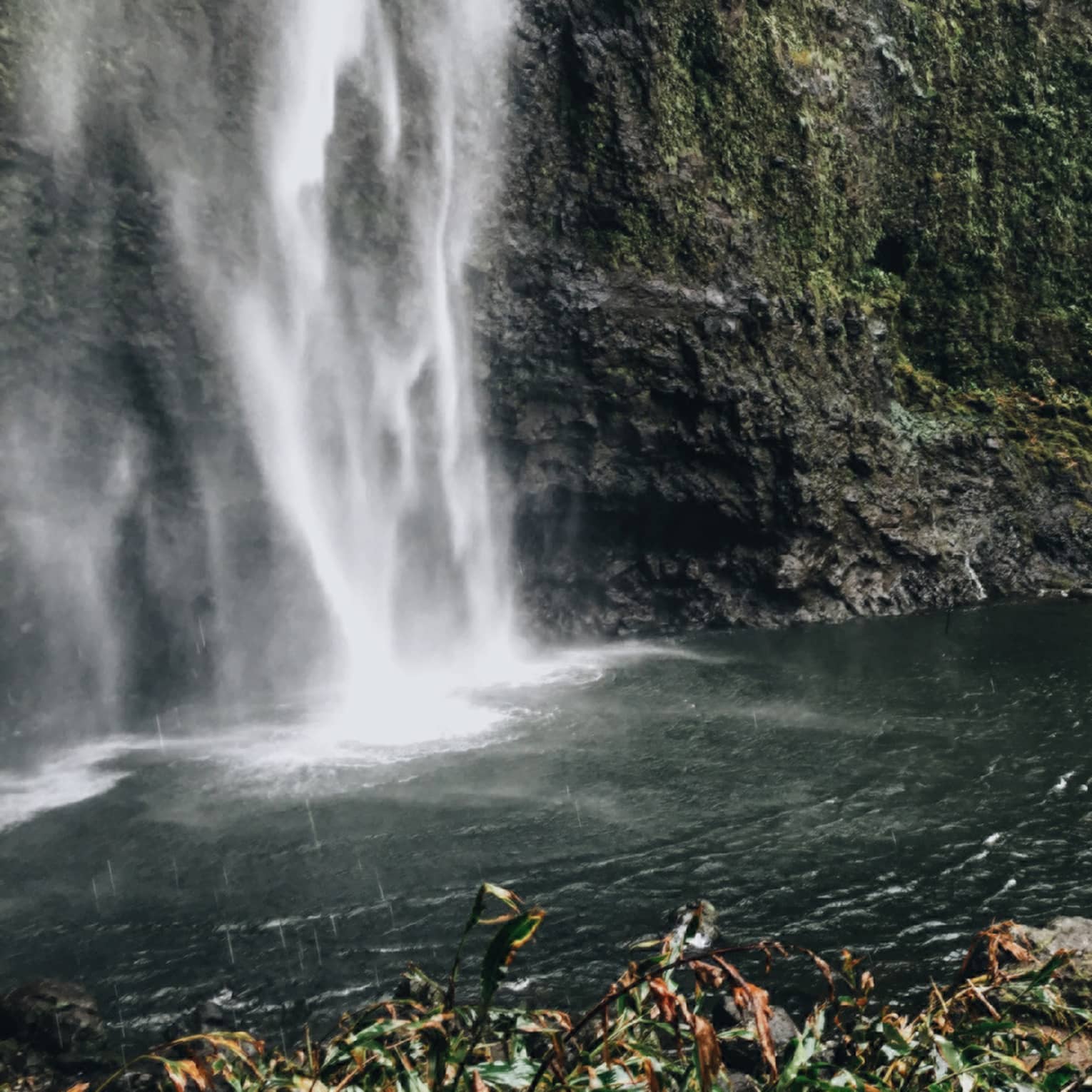 Back of woman hiking over black rocks under tall waterfall, small pond