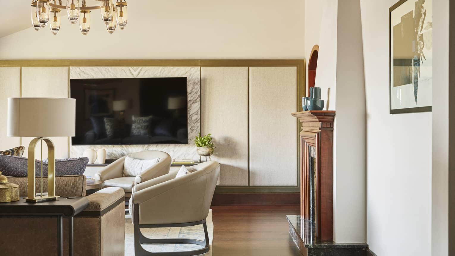 Hotel suite contemporary neutral living area under brass chandelier with flat-screen TV on one wall and wooden fireplace on another