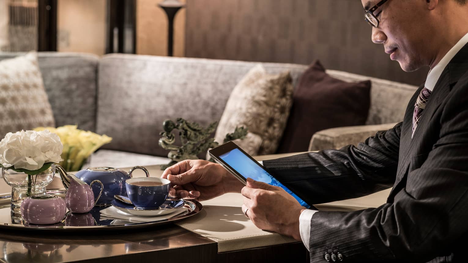 Man in suit holds tea cup on tray, reads iPad by elegant purple sofa