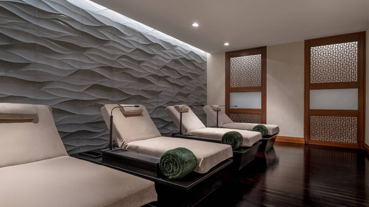 Spa relaxtaion room with a row of four upholstered lounge chairs in natural linen, each with a green rolled up blanket, a head pillow and a small lamp, all set in front of a textured wall