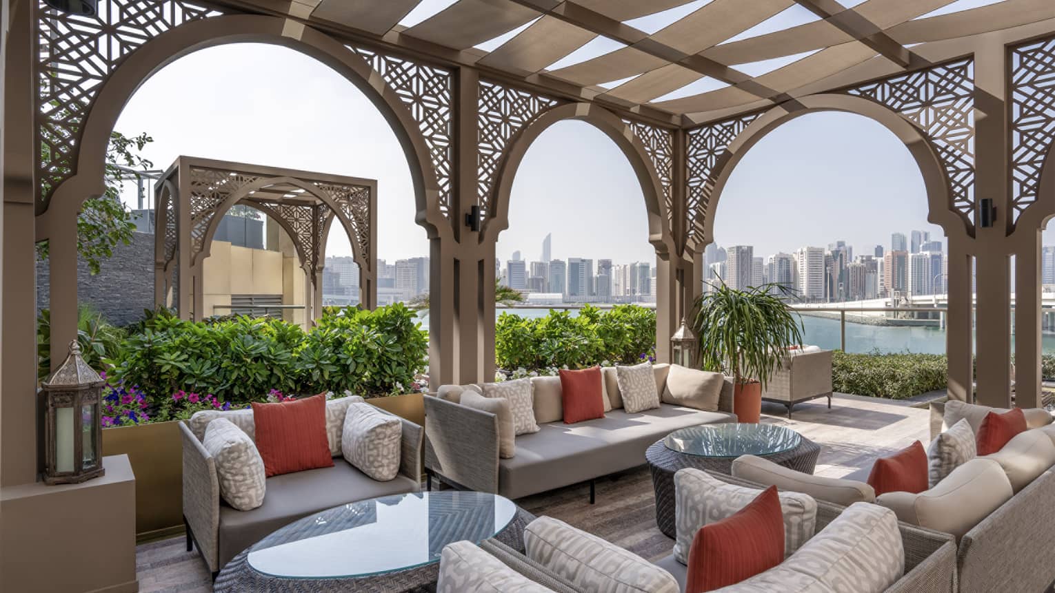 Outdoor terrace with light-grey lounge seating set beneath an Arabian-style pergola with a city skyline seen in the background