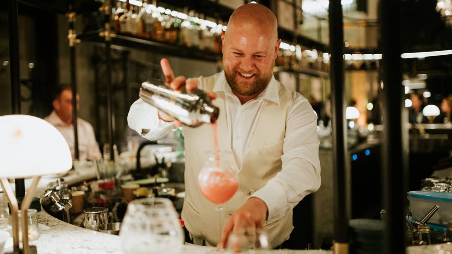 Bartender behind bar pouring a pink drink from a shaker into a tall glass