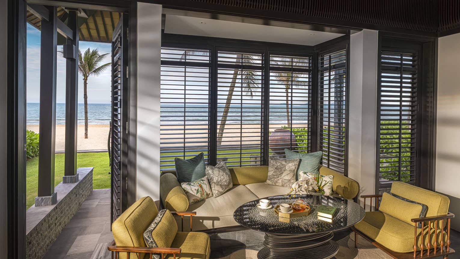 Guest room living area with mustard-coloured arm chairs, round coffee table and floor-to-ceiling windows with beach views