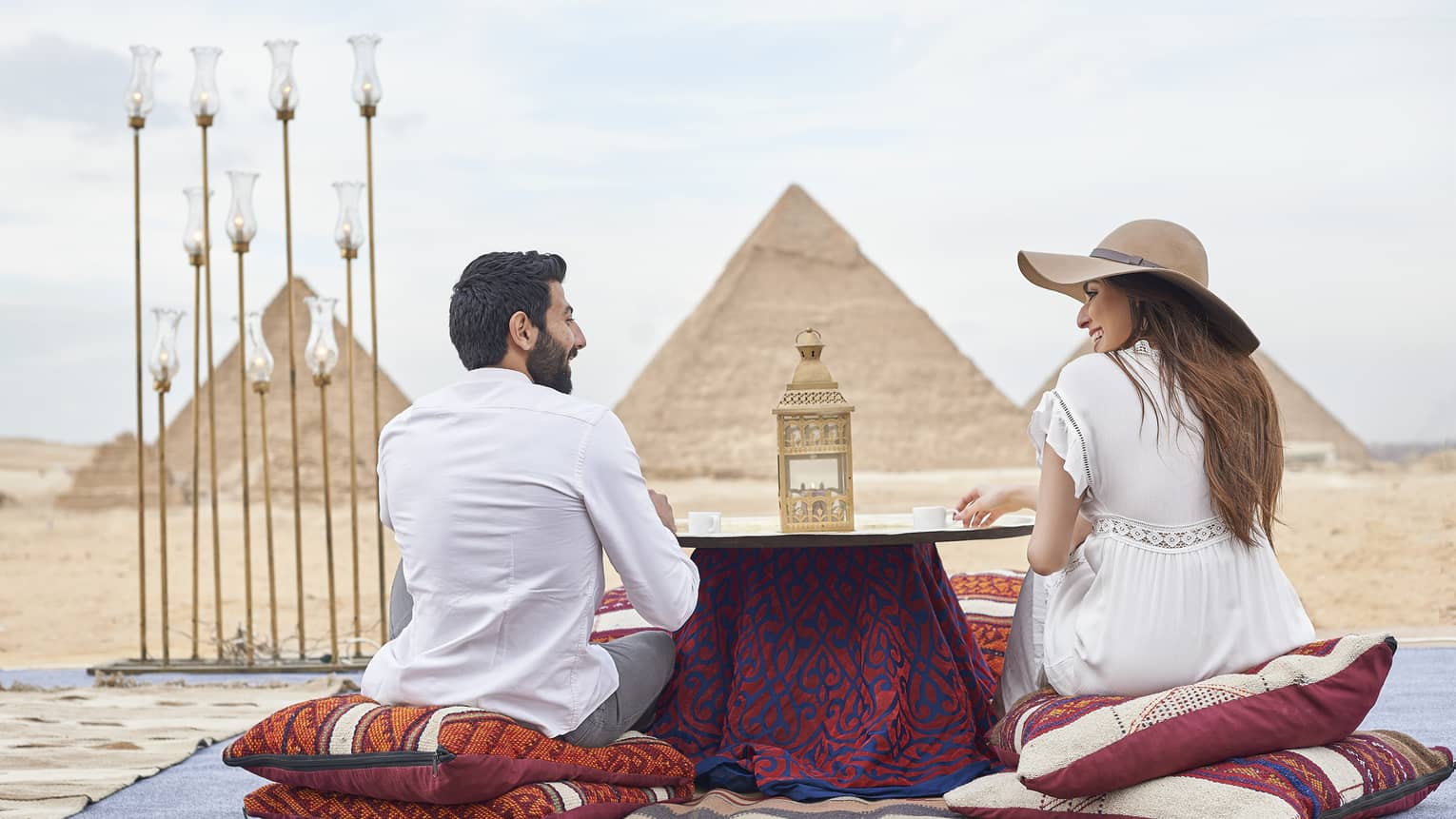 Back of couple sitting on pillows by small dining table in desert looking at pyramids