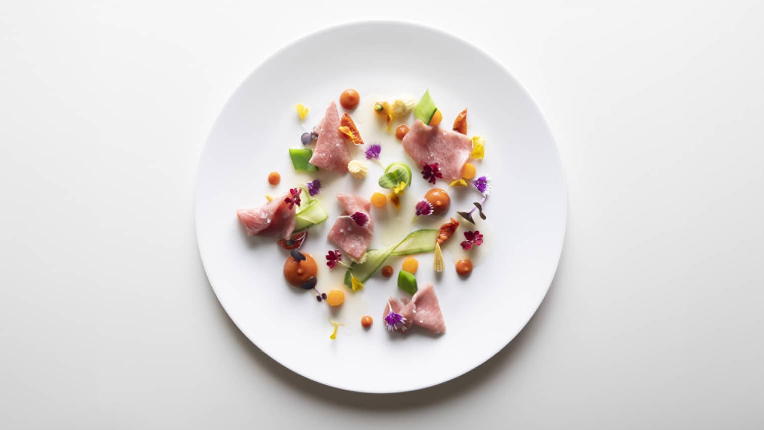 Slices of protein artfully arranged with dollops of brightly coloured sauce and sprigs of greenery and flowers on a white plate