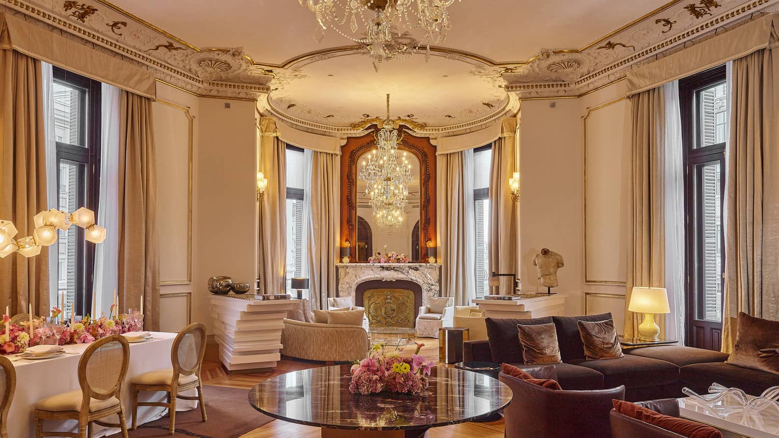 Large Regency-style living and dining area with tall ceilings, ornate finishes, two seating areas, a rectangular dining table and four sets of black-framed, floor-to-ceiling windows with beige curtains pulled to the side