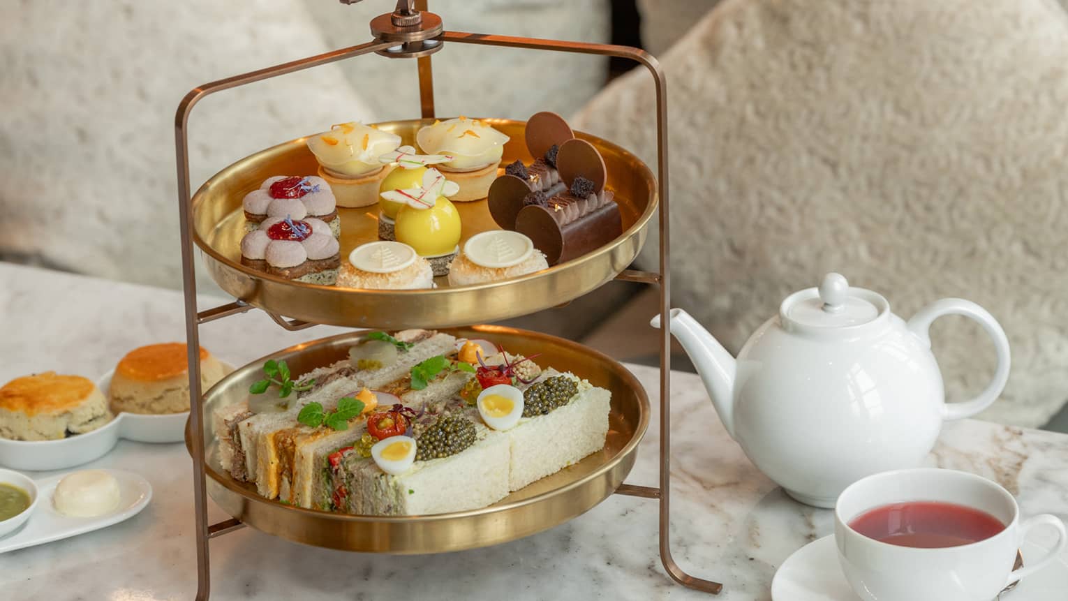 Two-tiered afternoon tea spread on marble coffee table in front of tan sofa