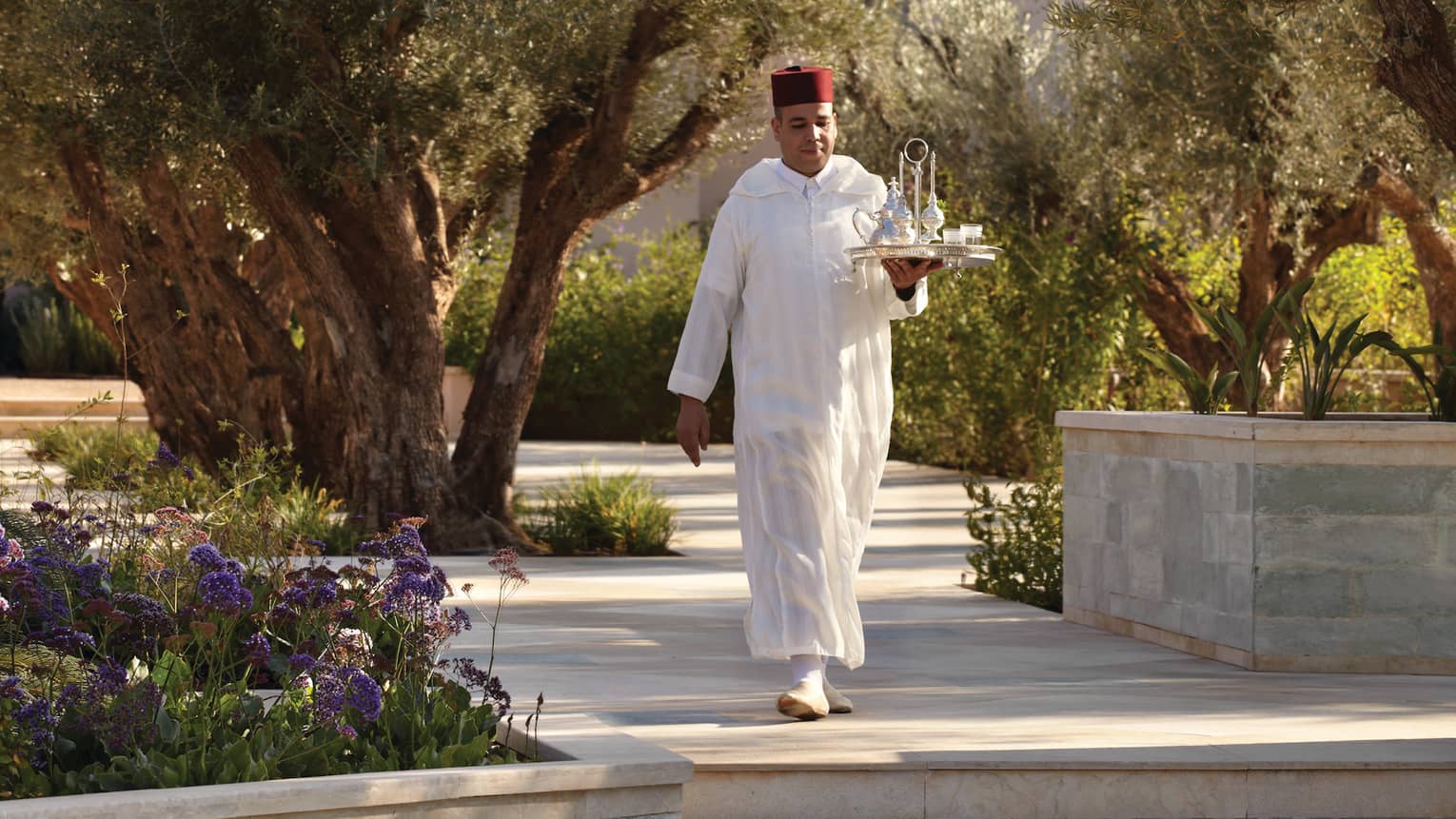 Server carries tray with silver teapots down sunny pathway