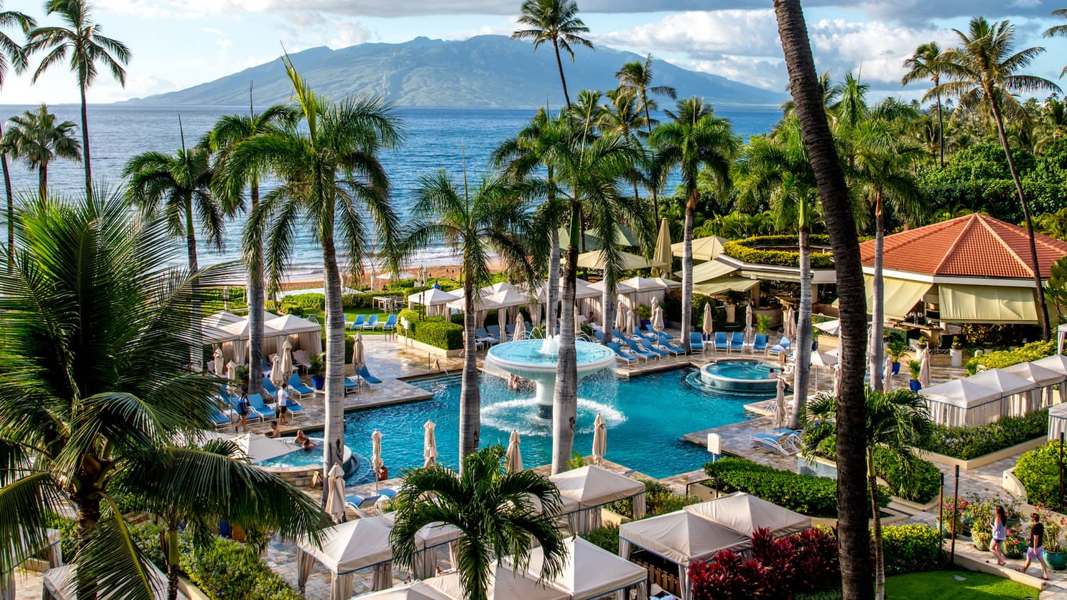 Aerial view of fountain in pool, surrounded by cabanas, palm trees, mountains