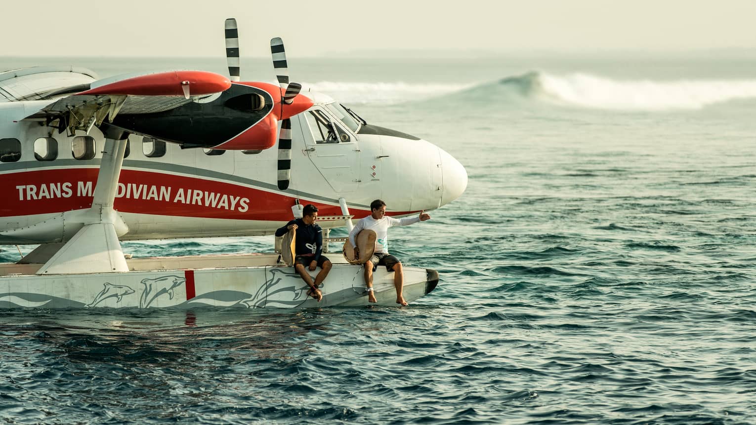 Two people holding surfboards sit on edge of float plane over water