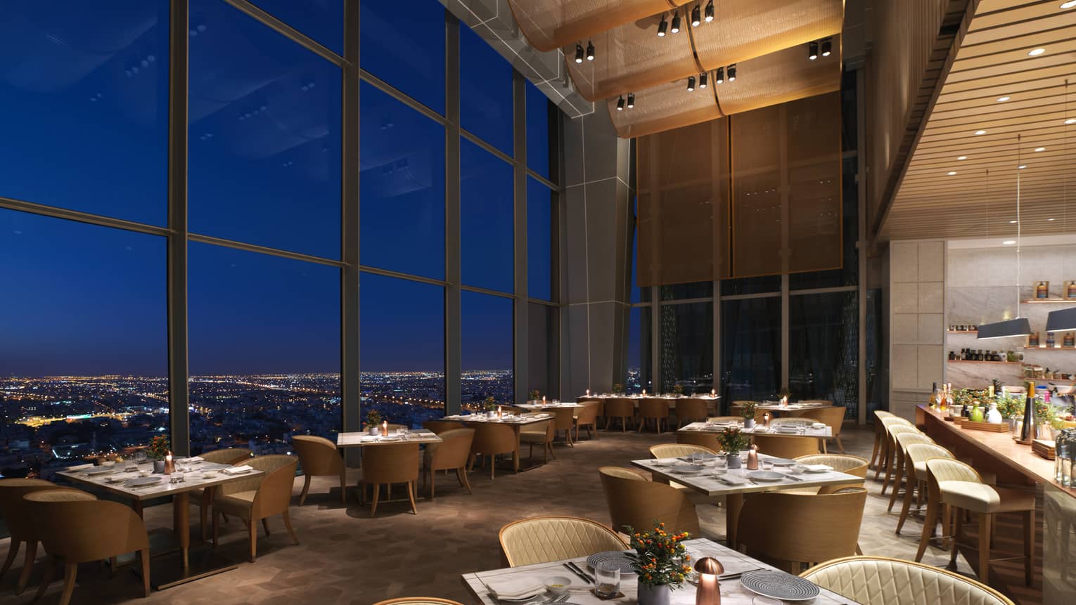 A dimly lit room with with a series of dining tables set up next to floor to ceiling windows.