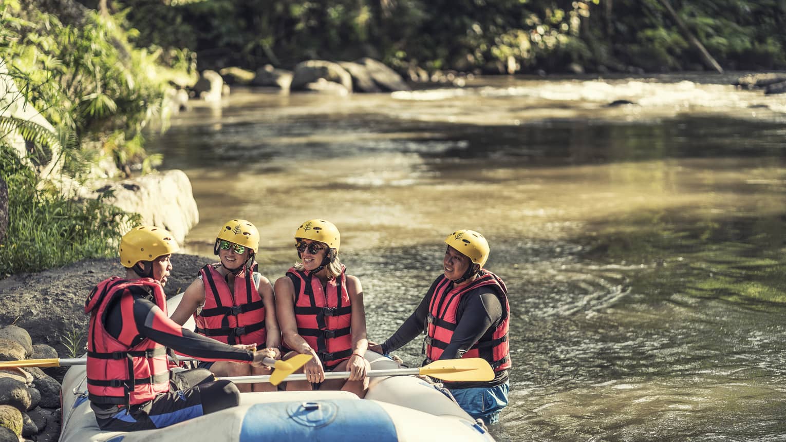 Four people wearing red life jackets, sunglasses and yellow helmets sit facing each other in a river raft