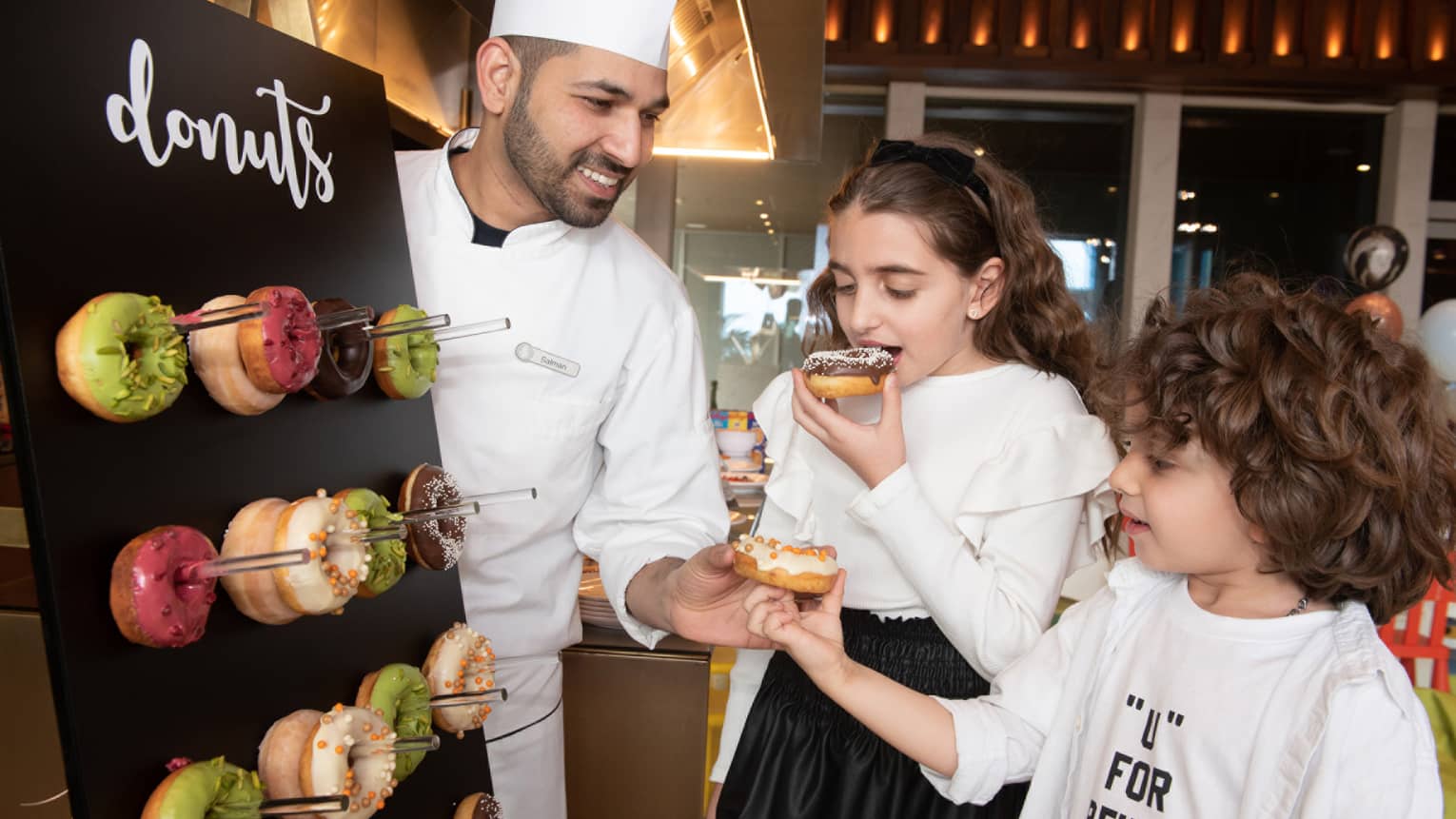 A chef wearing a white chef's jacket, apron and tall white hat hand two children donuts from a donut board
