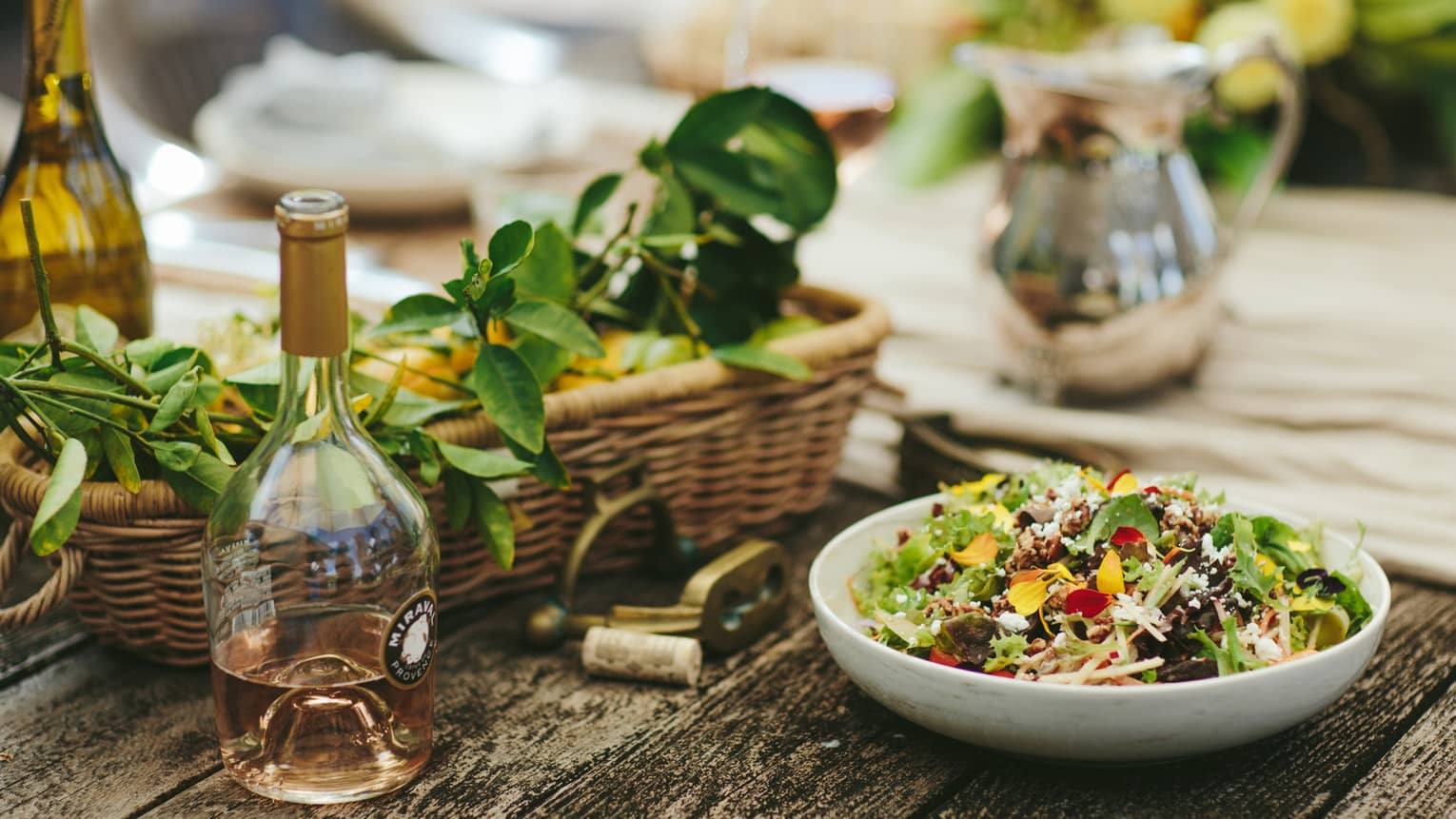 Bowl of salad next to wicker basket filled with leaves, bottles of wine on wooden table