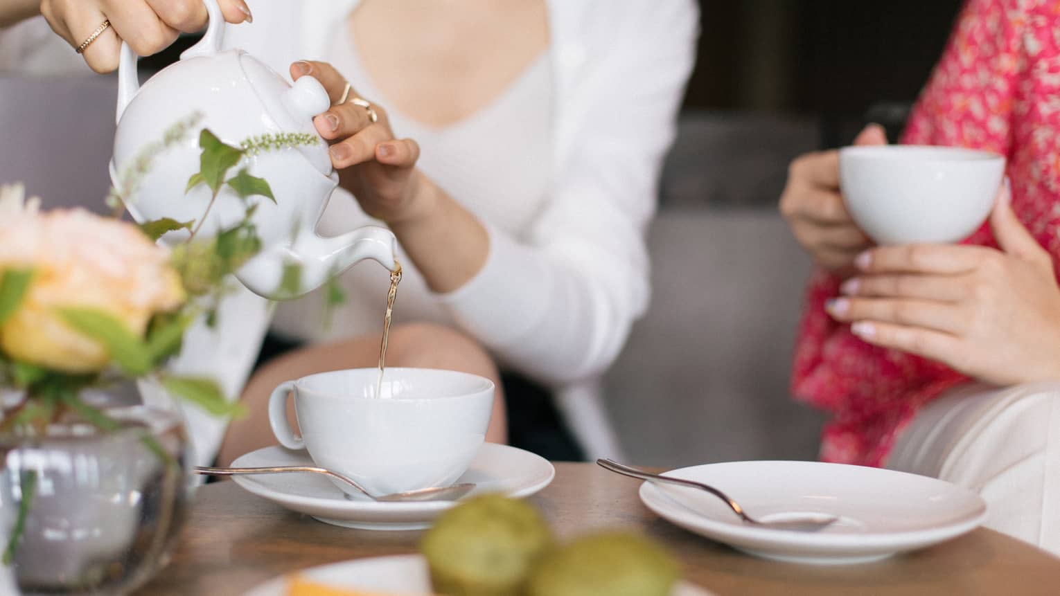 Two people sit at a table set for tea, with cups, saucers, a plate of small food items and a floral arrangement. One person is pouring tea from a teapot into one of the teacups.
