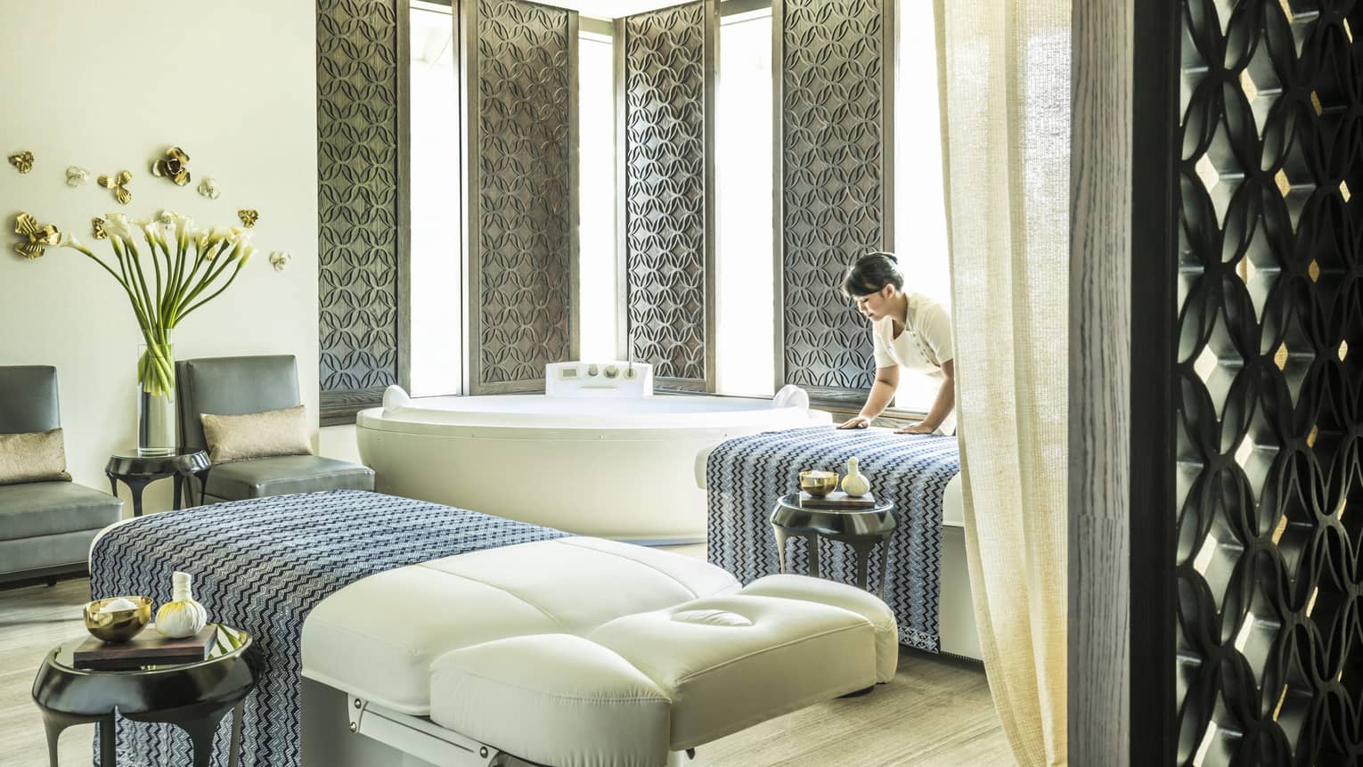 Large white spa table in front of white spa tub, silver wall panels, woman places blanket on table