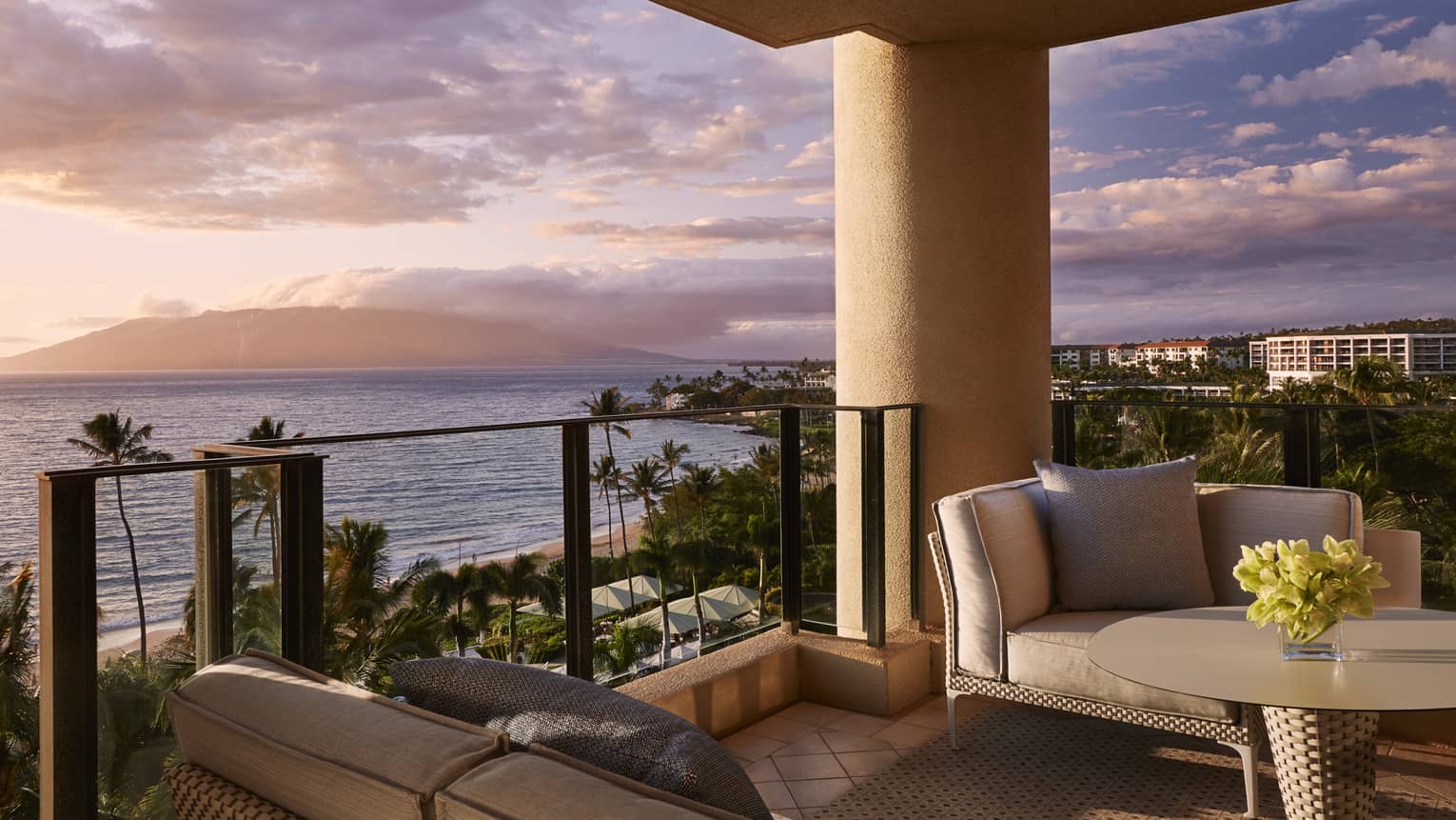 Balcony with cushioned seating and table overlooking beach palm trees and ocean at sunset